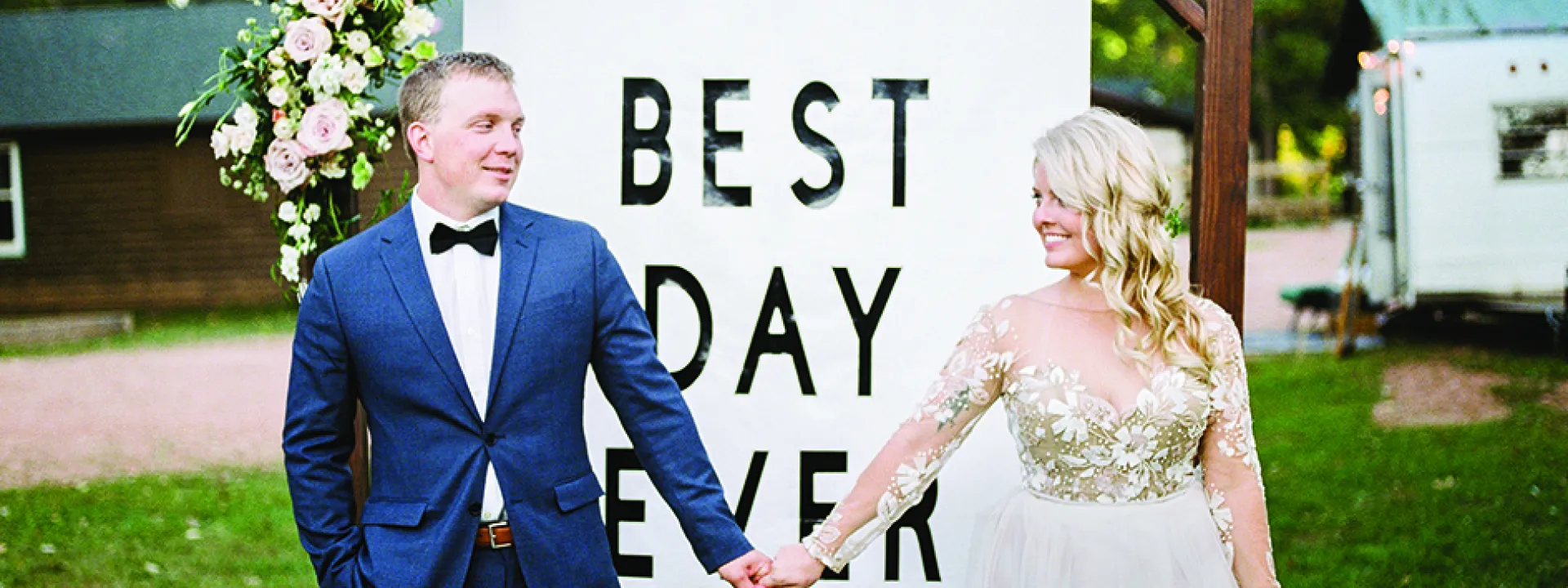 Ashley Zelenka and Evan Bohman hold hands in front of a Best Day Ever sign at their rustic camp wedding.