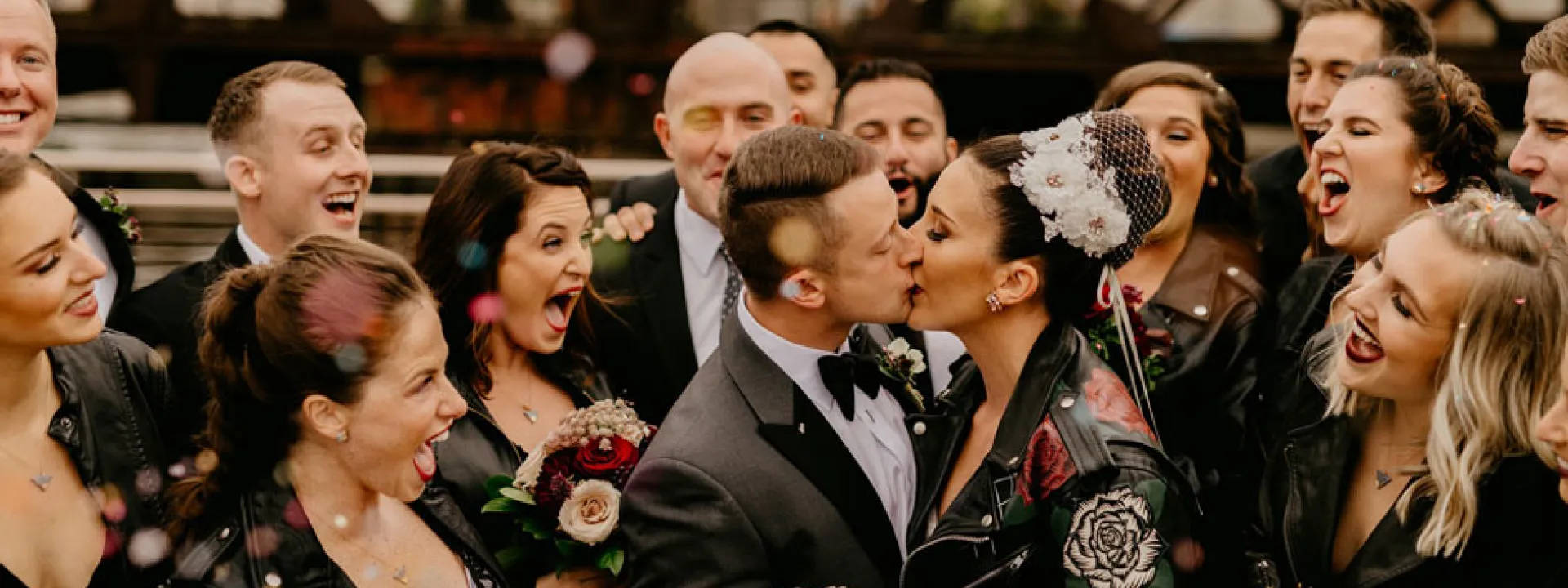 The bridal party looks on as the couple kisses near a bridge