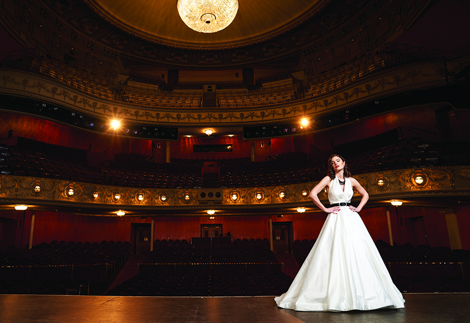 A bride stands on stage modeling a Legends Romona Keveza style No. L7171 silk shantung taffeta ballgown with pleated halter from White Dress Bridal Boutique.