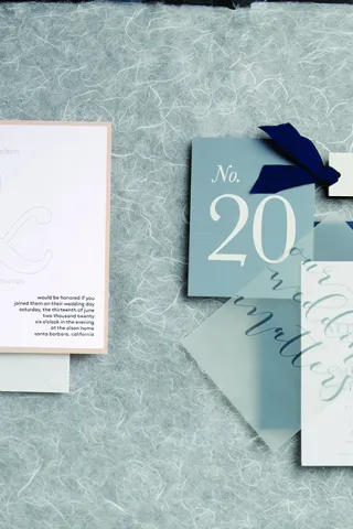 Two wedding invitations, one featuring embossed names and a marbled design and the other featuring a vellum overlay and the wedding hashtag