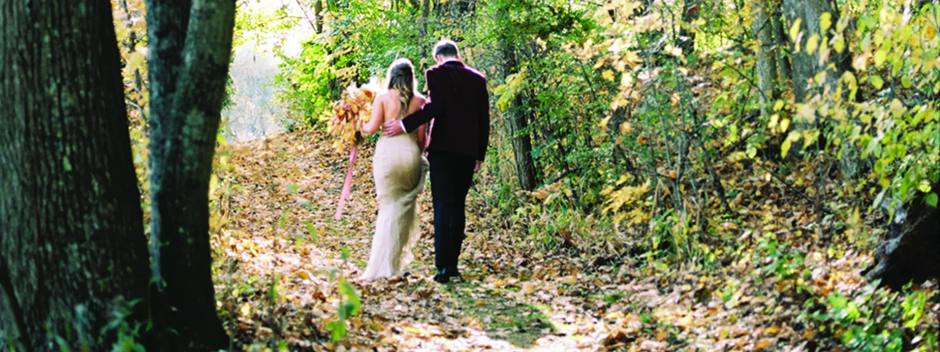 A bride and groom walk a path at the golden hour. His burgundy suit and her neutral wedding dress complement the fall wedding theme.